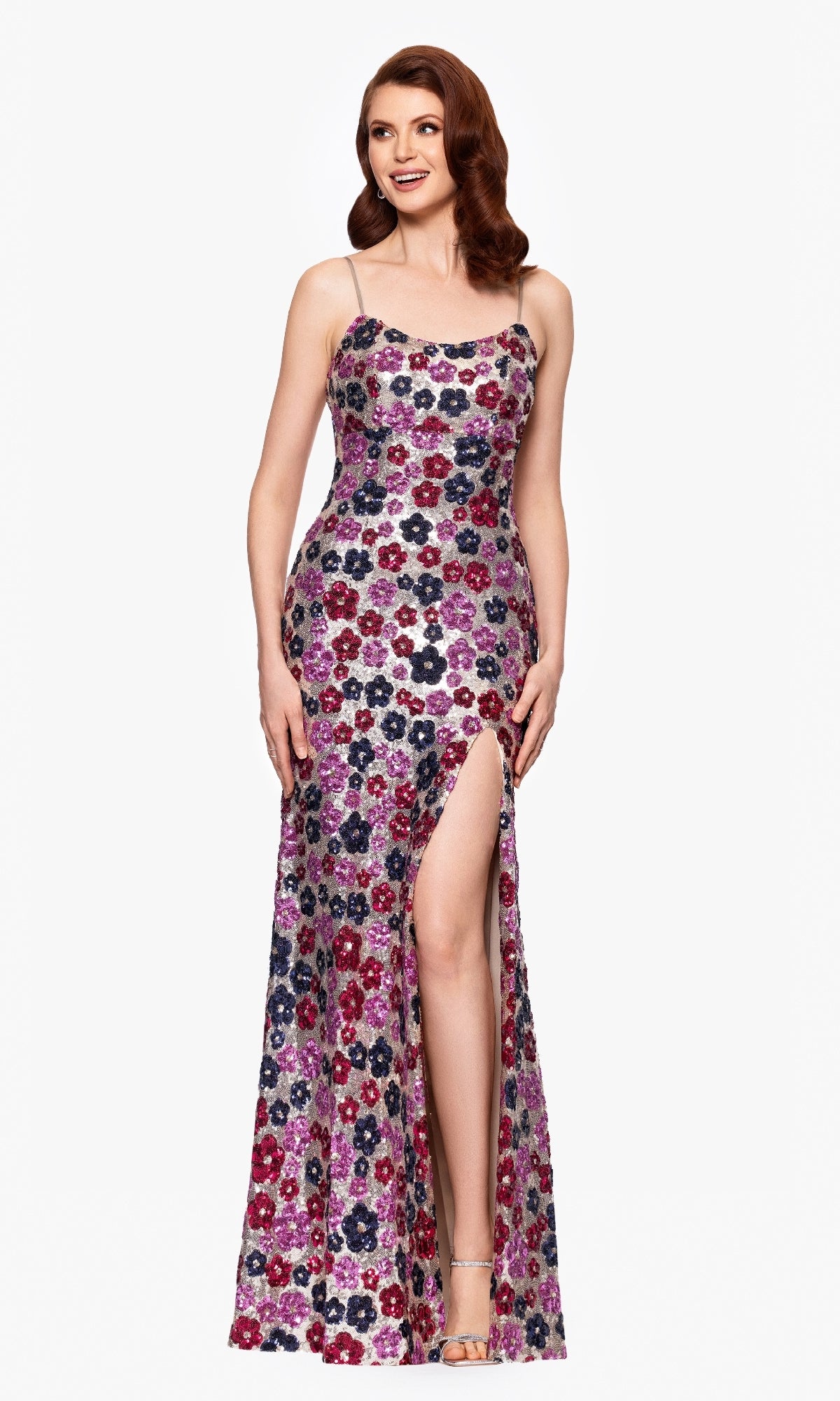 A woman stands and smiles, wearing the Betsy & Adam A27017 Floral-Sequin Long Prom Dress with colorful patterns, sleeveless design, floor length, and a thigh-high slit against a plain white background.